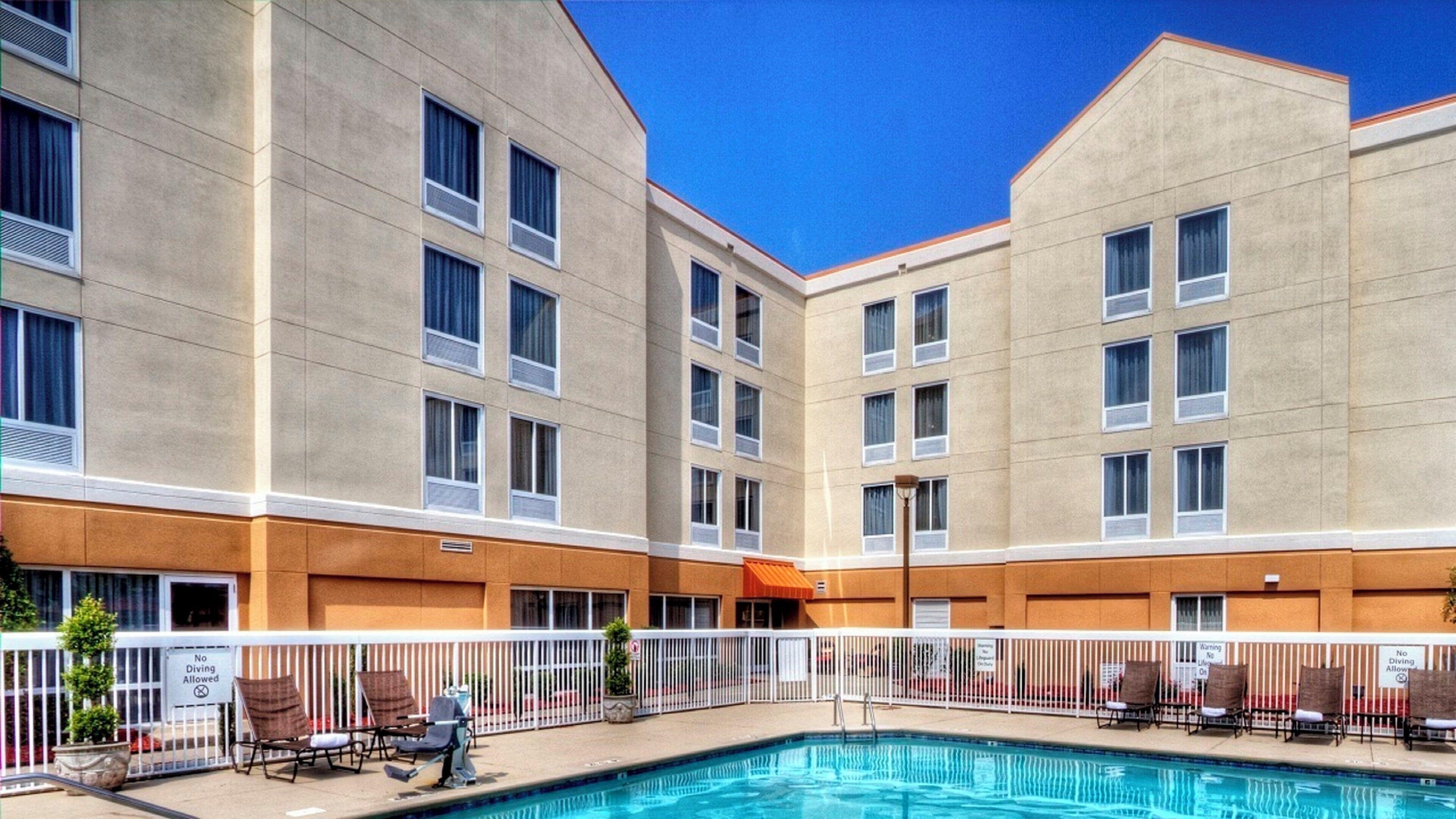 Un hotel con piscina, varios balcones y un área exterior vallada con sillas de descanso.