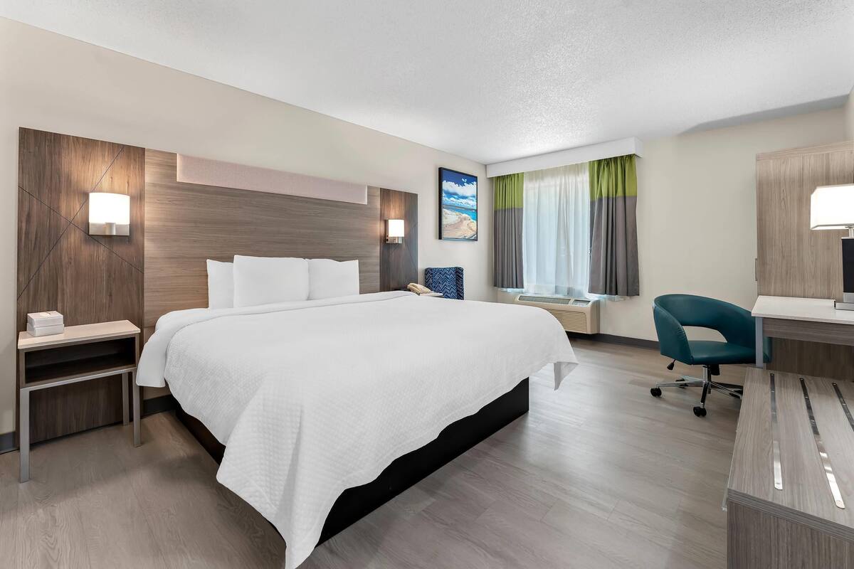 A hotel room with a large bed, bedside tables, a desk with a chair, a window with curtains, and a painting on the wall.
