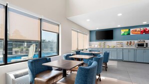 A modern dining area with blue upholstered chairs and wooden tables, a flat-screen TV mounted on the wall, and a view of a swimming pool through large windows. - Quality Inn Sandersville (Sandersville)