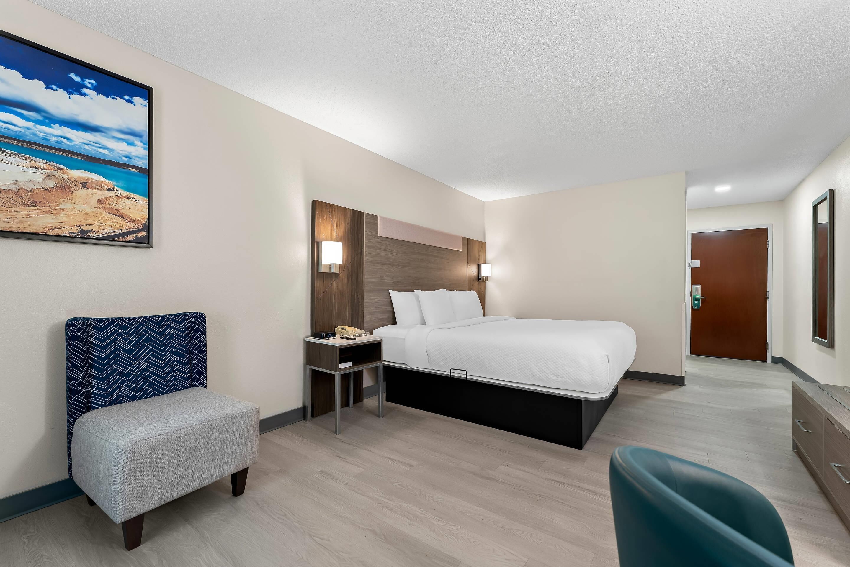 A hotel room with a bed, a nightstand, a chair, a desk, and a painting on the wall.