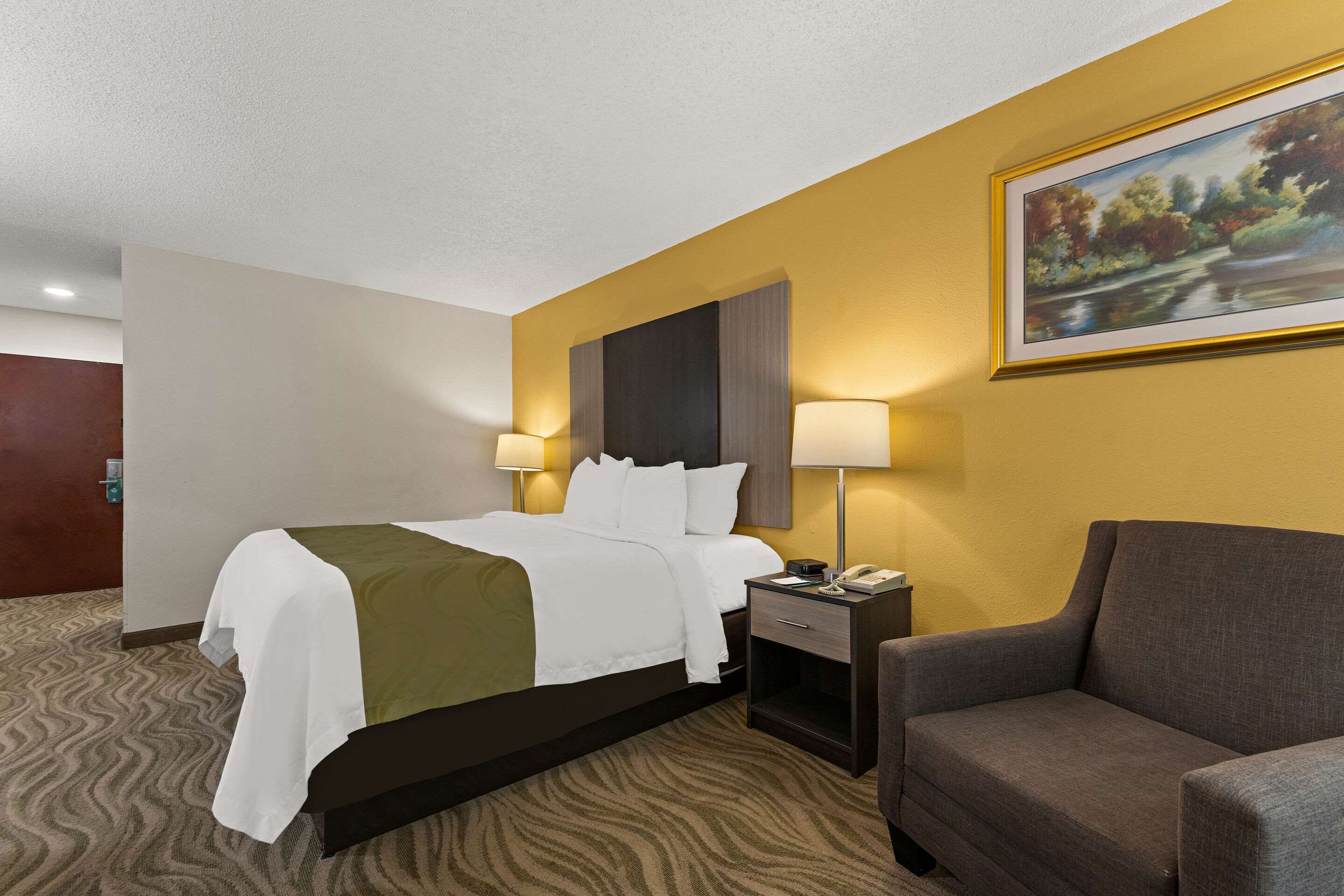 A hotel room with a bed, two bedside lamps, a nightstand, a chair, and a painting on the wall.