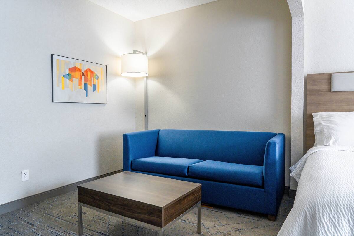 A hotel room with a blue sofa, a wooden coffee table, a white bed, and a painting on the wall.