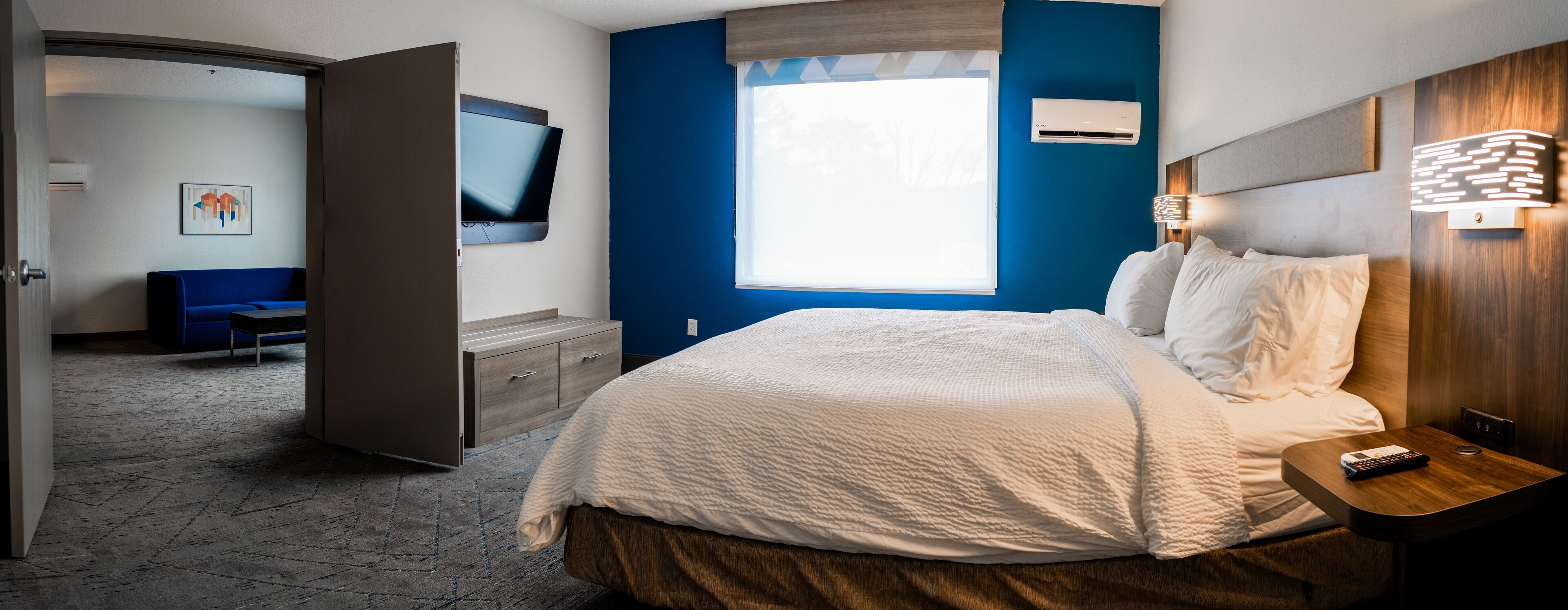 A modern hotel room with a large bed, a desk with a lamp, a TV, and a window with blinds.
