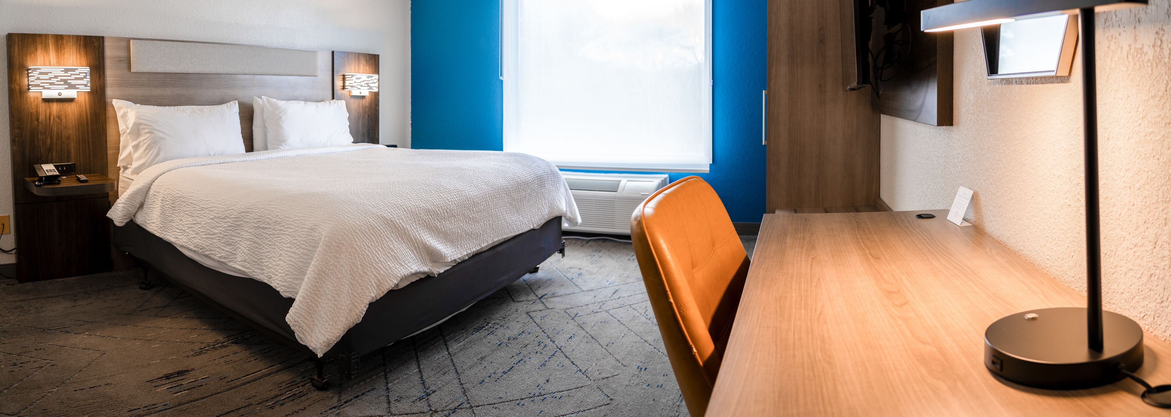 A hotel room with a bed, a desk, a chair, a nightstand, and a wall-mounted lamp.