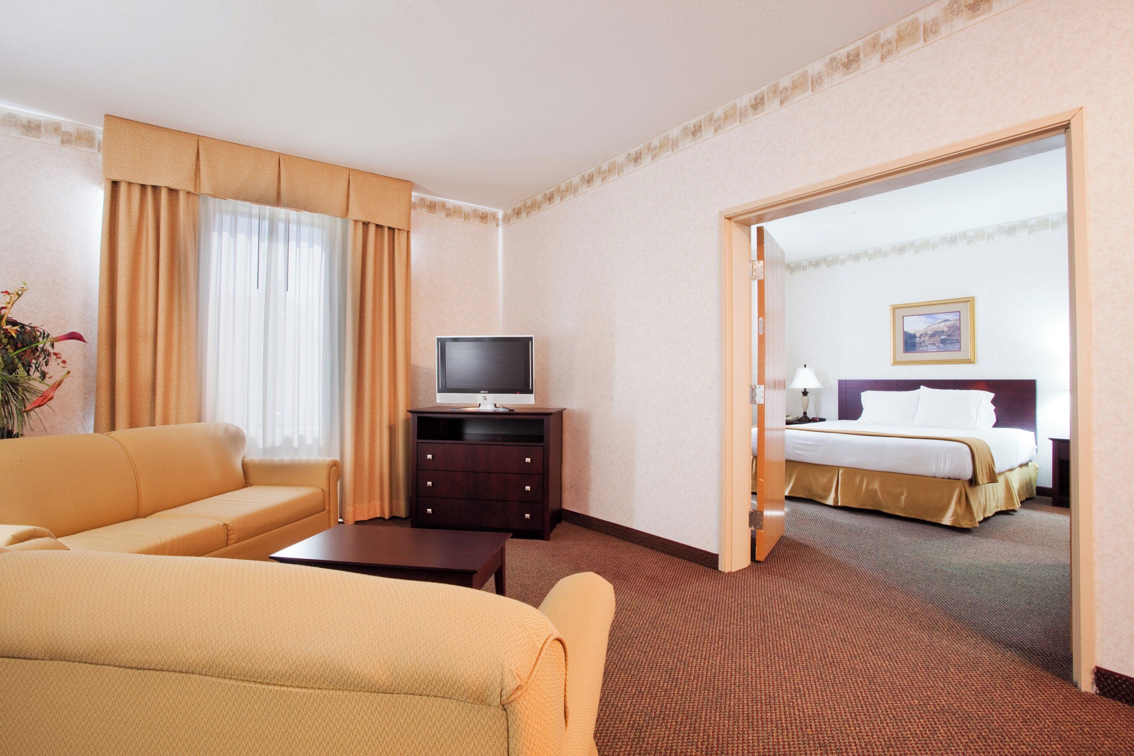 Habitación de hotel con sofá, televisor sobre un mueble bajo, cama y puerta que da a otra habitación.