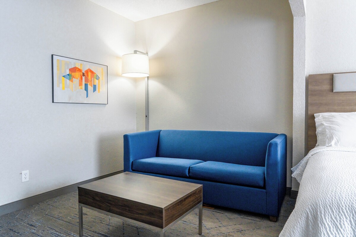 Une chambre d’hôtel avec un canapé bleu, une table basse en bois, un lit blanc et un tableau au mur.