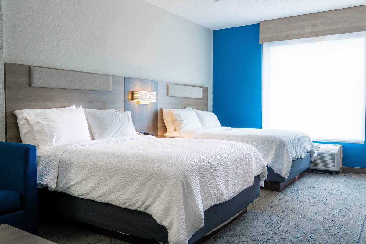 A hotel room with two beds, a blue accent wall, and a large window providing natural light.