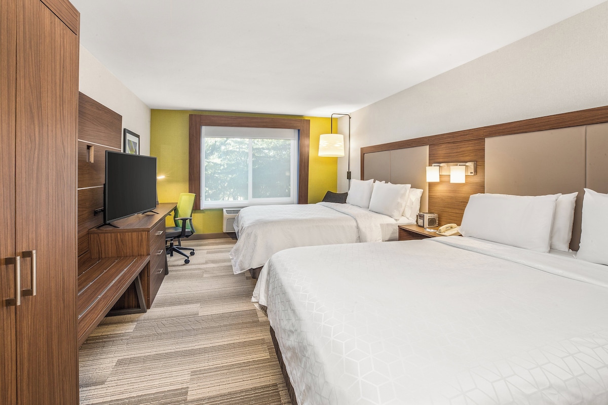 A hotel room with two beds, a desk with a television, a chair, and a window with a view of trees.