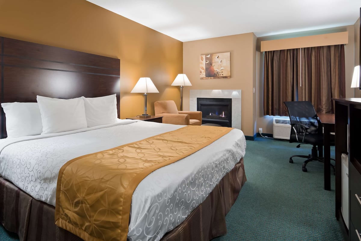 A hotel room with a large bed, two bedside lamps, a chair, a fireplace, a desk with a chair, and a television.