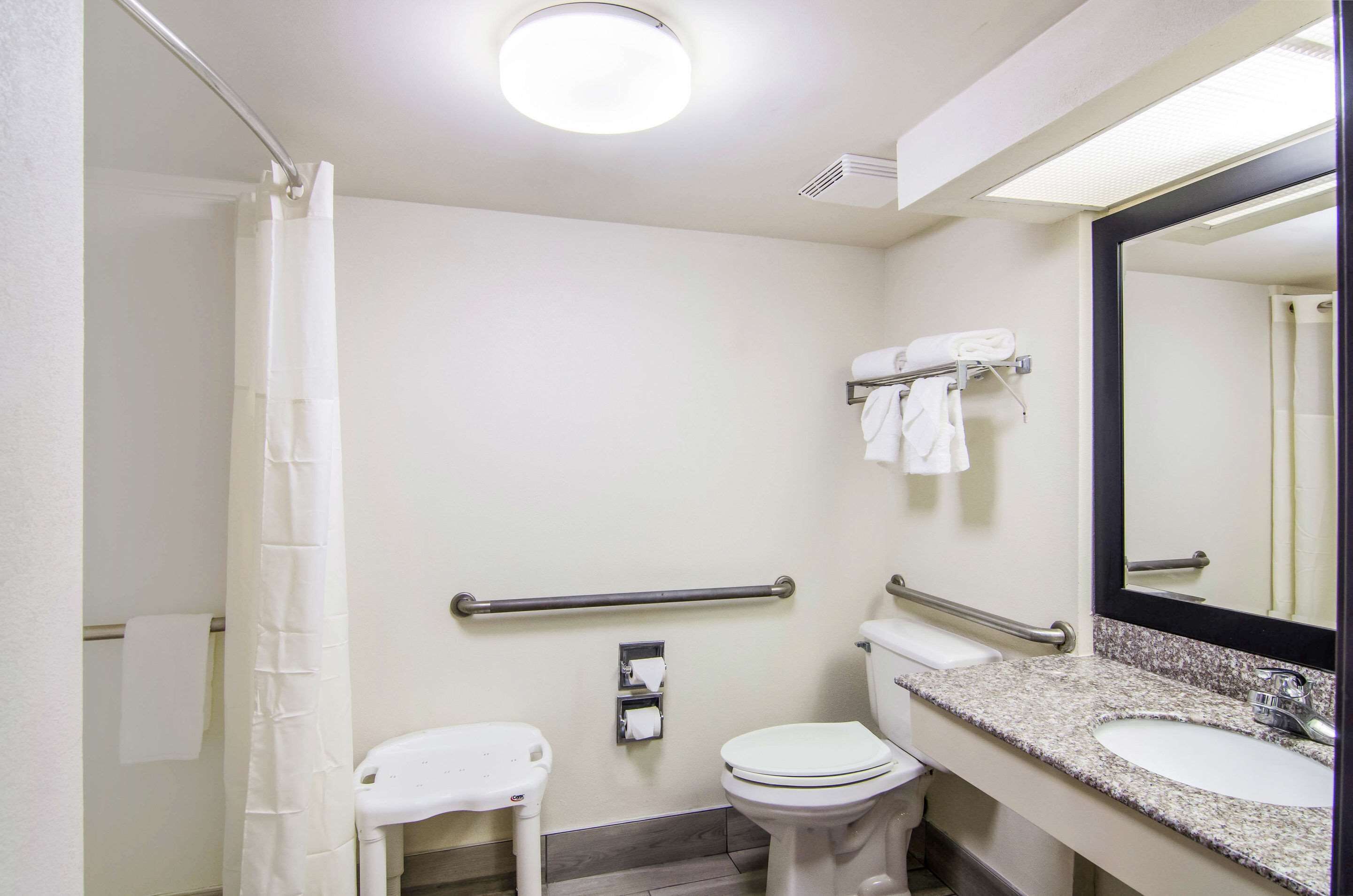 A small, clean bathroom with a toilet, sink, and shower. There are handrails and a mirror.