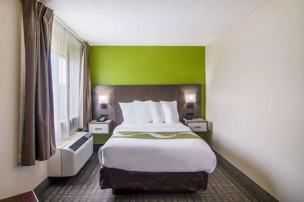 A hotel room with a bed, nightstands, a heater, and a window with curtains.