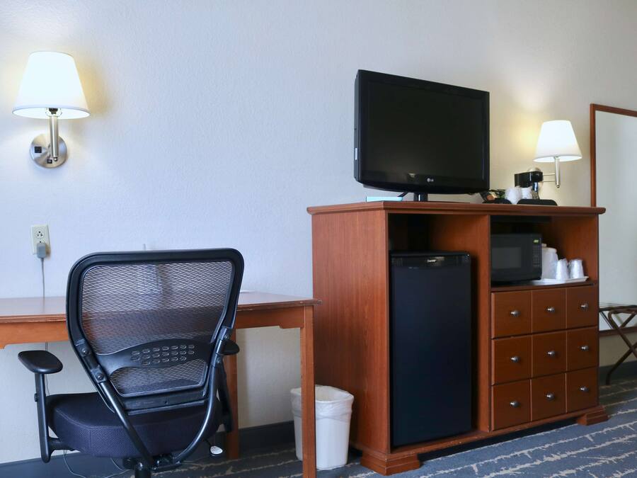 Une chambre d’hôtel équipée d’un bureau, d’un fauteuil de bureau noir, d’un téléviseur posé sur une commode en bois, d’un four à micro-ondes et d’une petite poubelle.