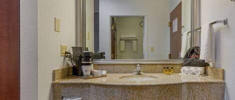 A bathroom with a granite countertop, a sink, a mirror, a towel rack, and a coffee maker.