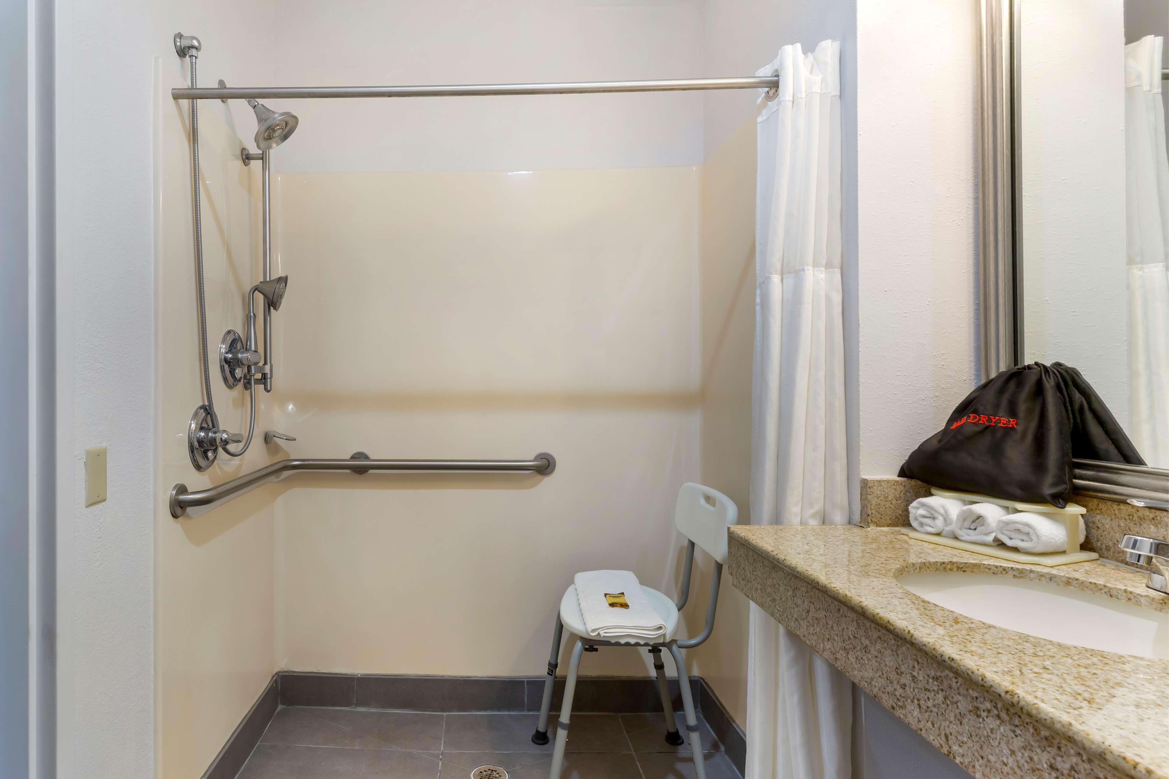 Un baño con ducha, barra de apoyo, una silla blanca, lavamanos y un toallero con toallas.