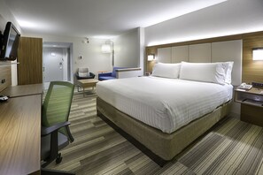 Quarto de hotel com uma cama grande, uma escrivaninha com cadeira, televisão e mesa de cabeceira com abajur.