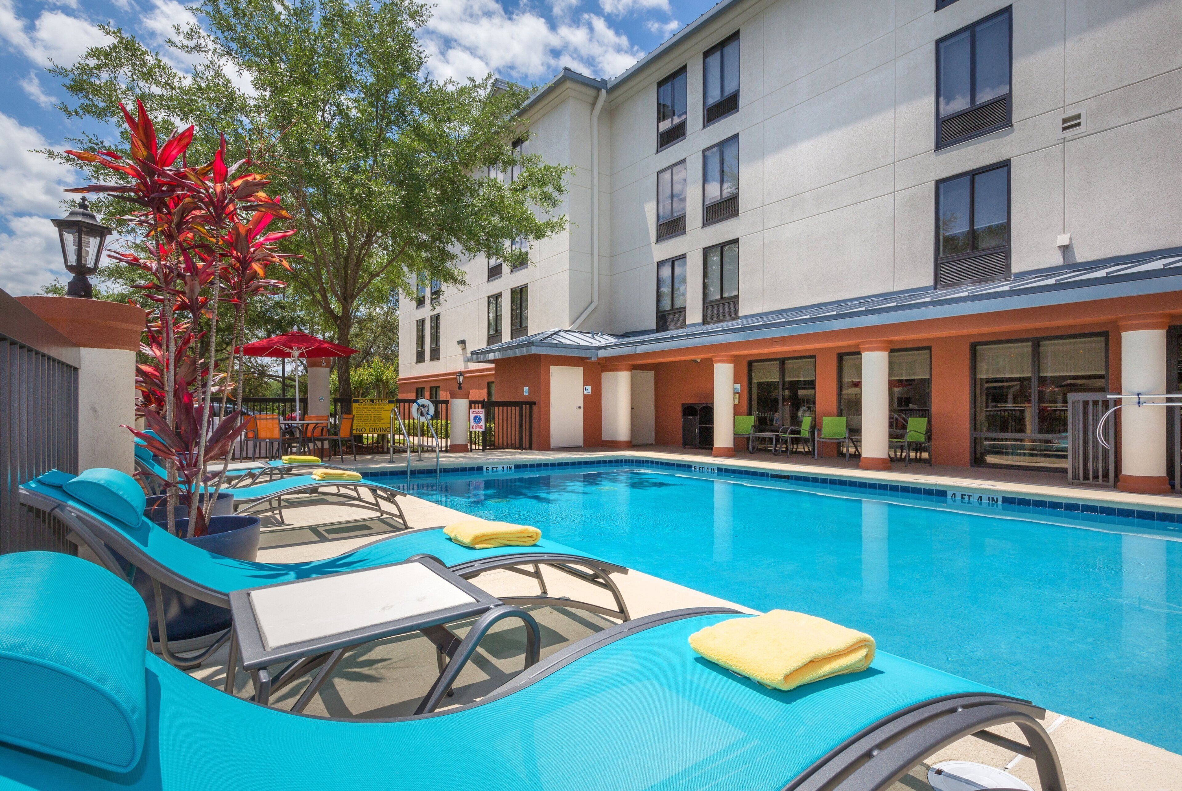A swimming pool with lounge chairs and umbrellas, surrounded by a building with multiple windows and a covered patio area.