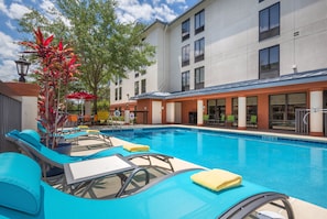 A swimming pool with lounge chairs and umbrellas, surrounded by a building with multiple windows and a covered patio area.