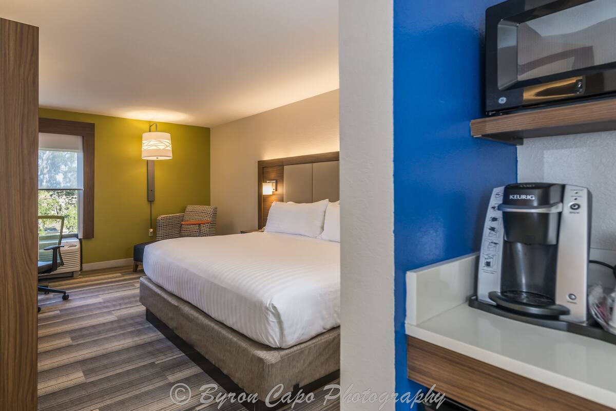 Una habitació d'hotel amb un llit, una taula de nit amb un fabricant de cafè, una cadira i una làmpada.
