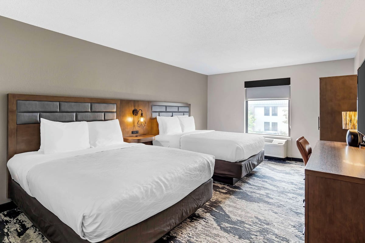 Une chambre d’hôtel avec deux lits, un bureau, une chaise, une table de chevet avec une lampe et une fenêtre donnant sur un paysage agréable.