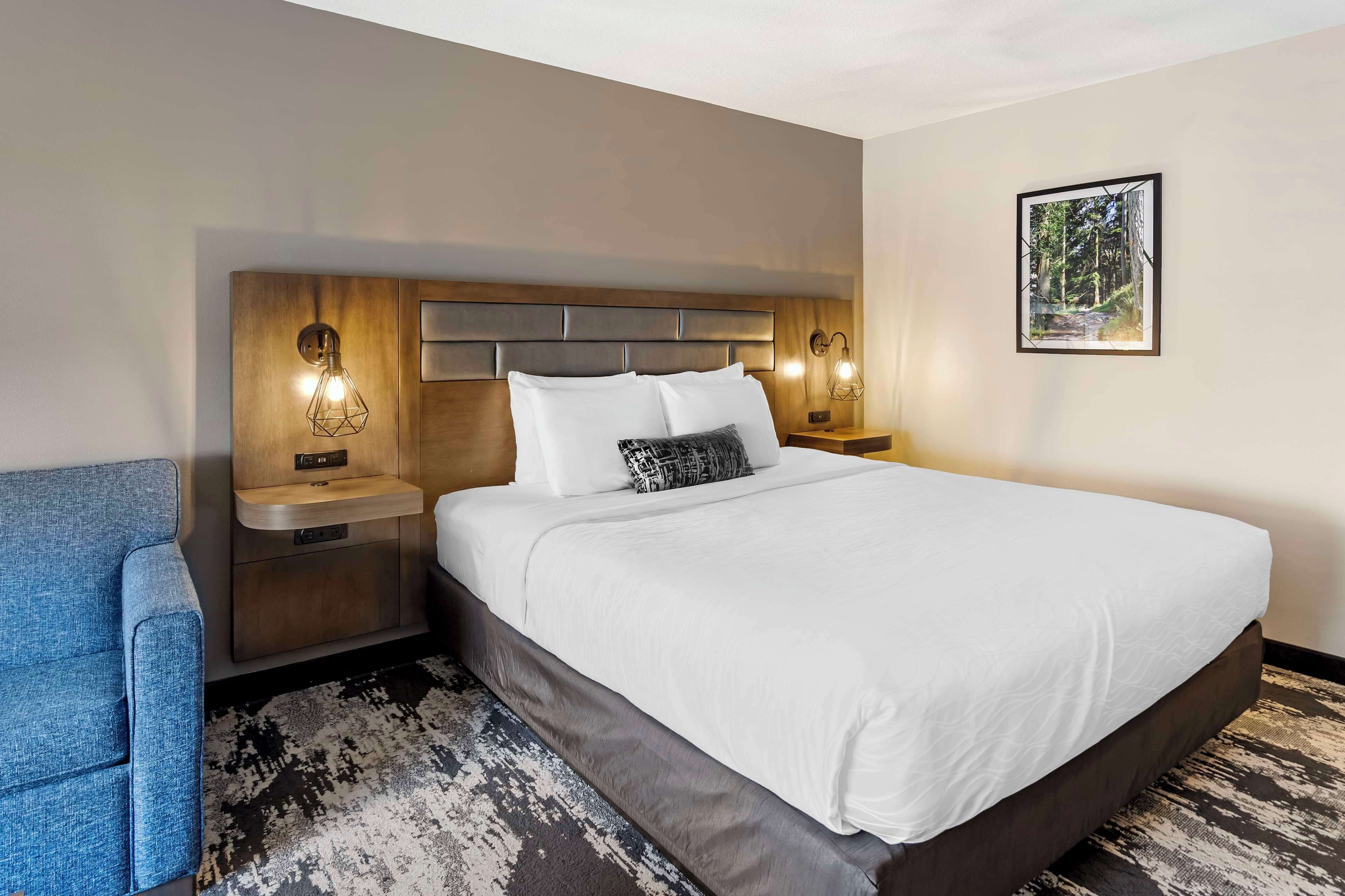A hotel room with a large bed, two bedside tables with lamps, a blue armchair, and a framed picture on the wall.
