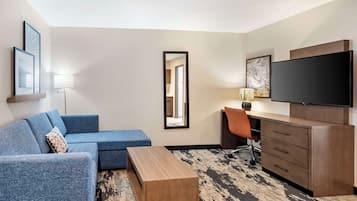 A modern hotel room with a blue sofa, a wooden coffee table, a desk with a chair, a flat-screen TV, and a large mirror.
