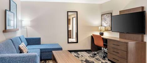 Ein modernes Hotelzimmer mit einem blauen Sofa, einem Holztisch, einem Schreibtisch mit Stuhl, einem Flachbildfernseher und einem großen Spiegel.