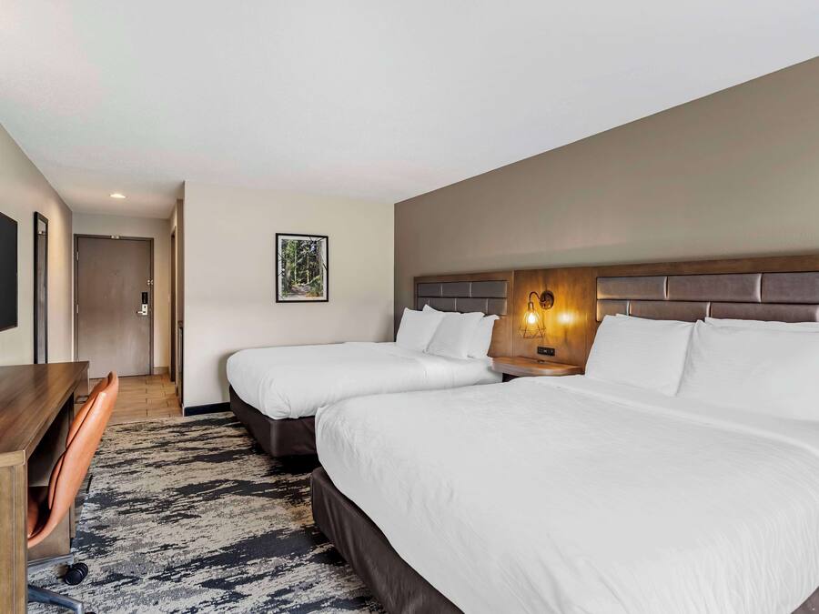 Quarto de hotel com duas camas, uma escrivaninha, uma cadeira, uma TV e um quadro na parede.