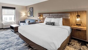 A hotel room with a large bed, a sofa, a bedside table, a lamp, and a window with a view of buildings. - Best Western Plus Bourbonnais Hotel & Suites (Bourbonnais)