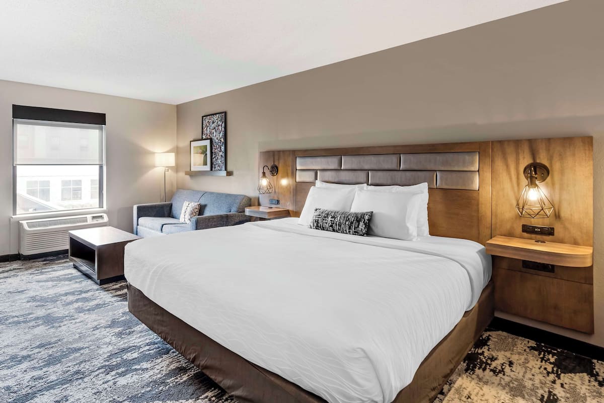 Une chambre d’hôtel avec un grand lit, un canapé, une table de chevet, une lampe et une fenêtre donnant sur des immeubles.