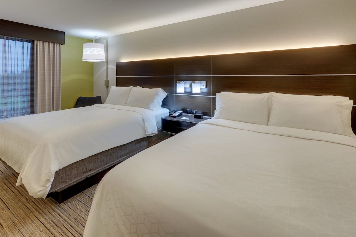 A hotel room with two beds, a bedside table with a lamp, and a wall-mounted control panel.