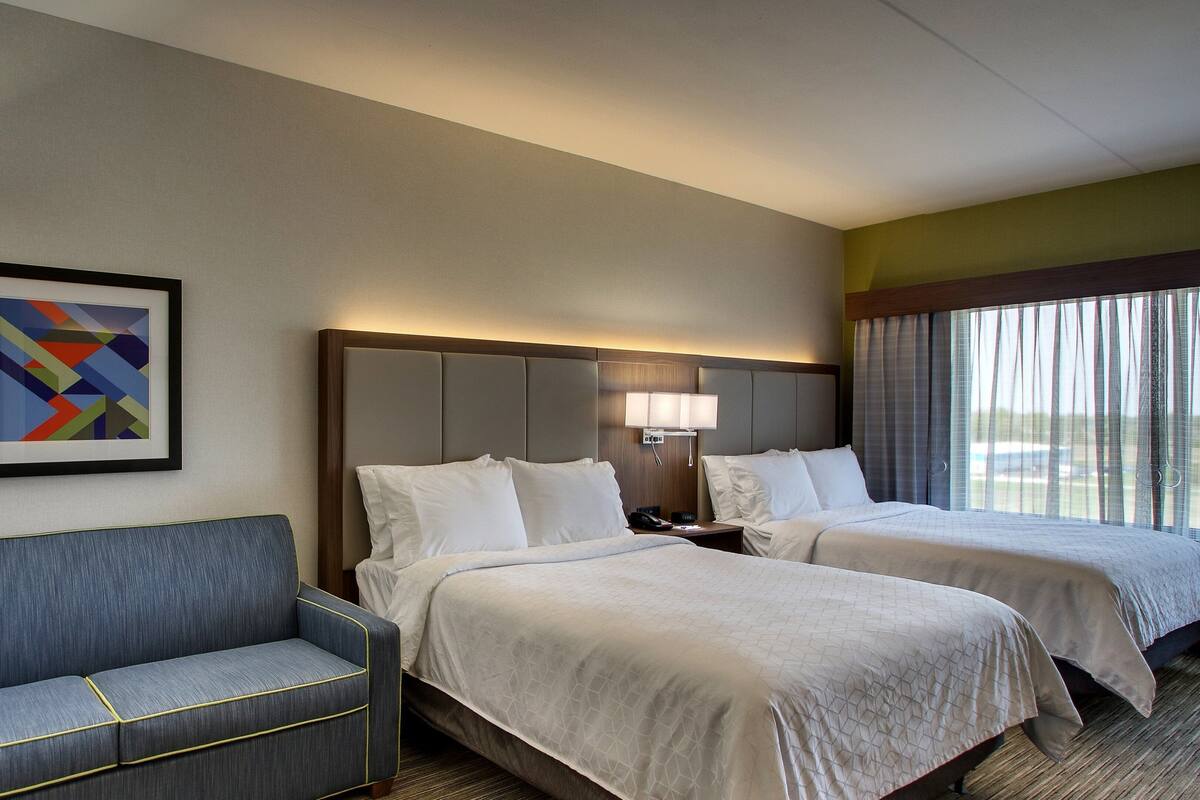 A hotel room with two beds, a sofa, a framed abstract painting, and a window with curtains.