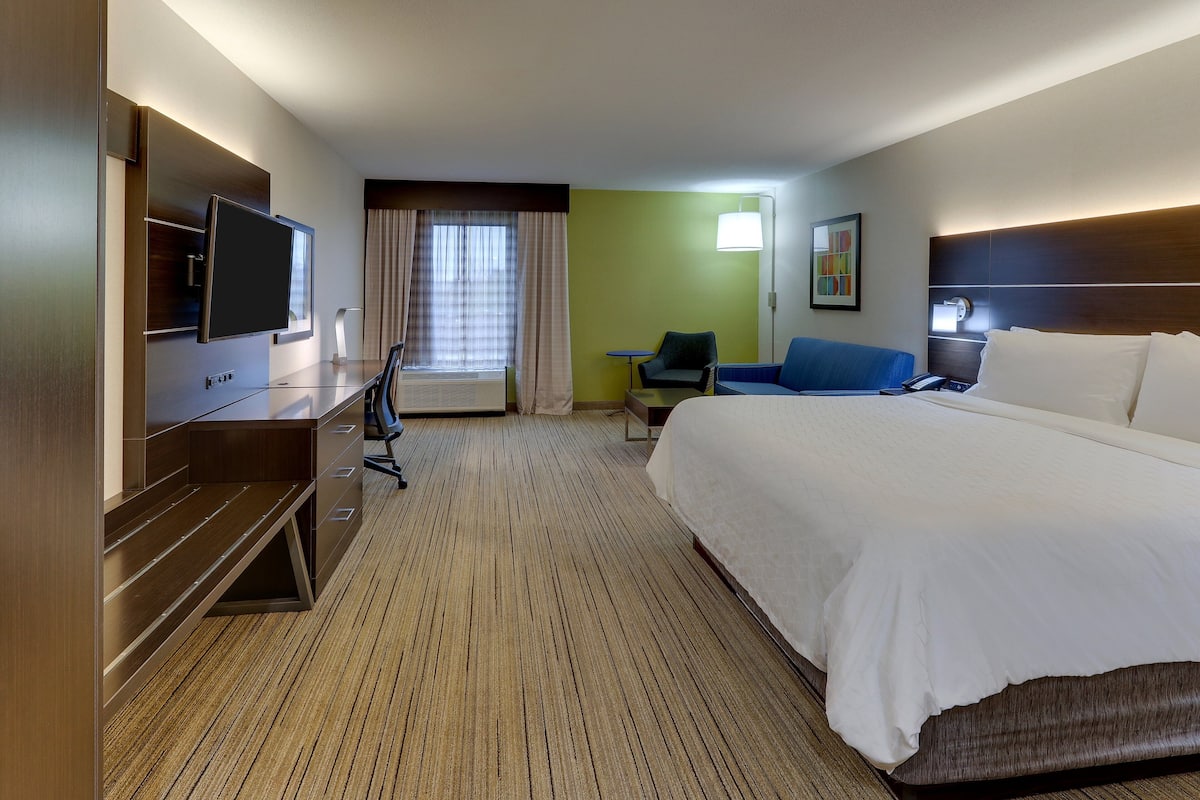 A hotel room with a large bed, a desk with a flat-screen TV, a chair, a small table, and a lamp.