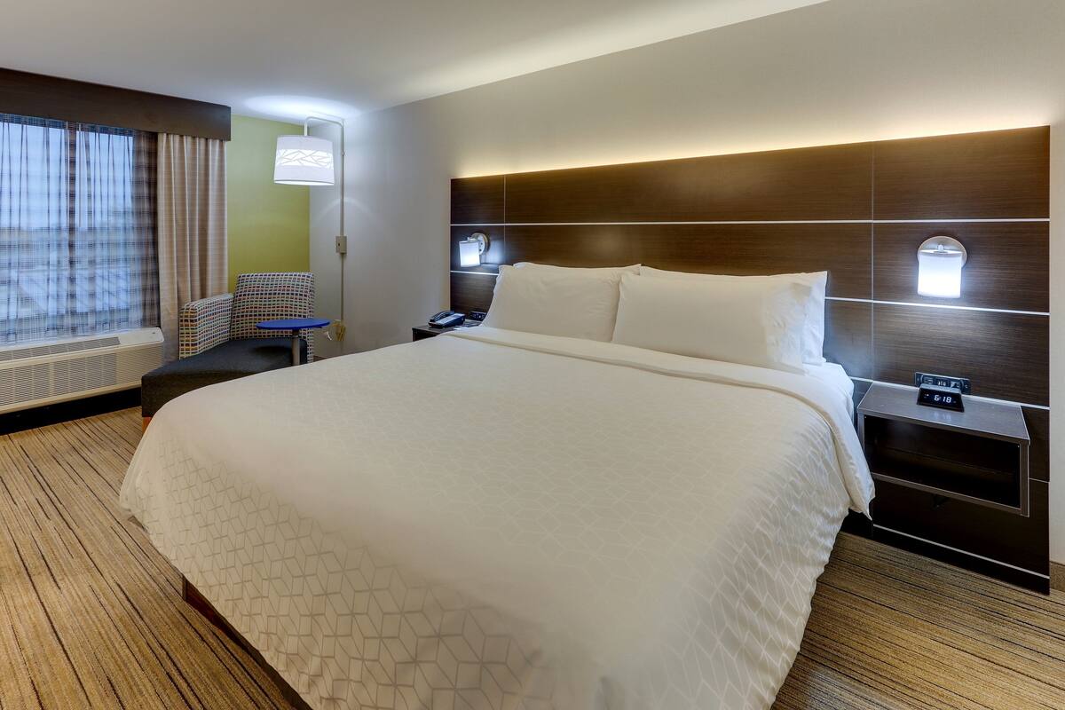 A hotel room with a large bed, a nightstand, a chair, a desk, a lamp, and a window with curtains.