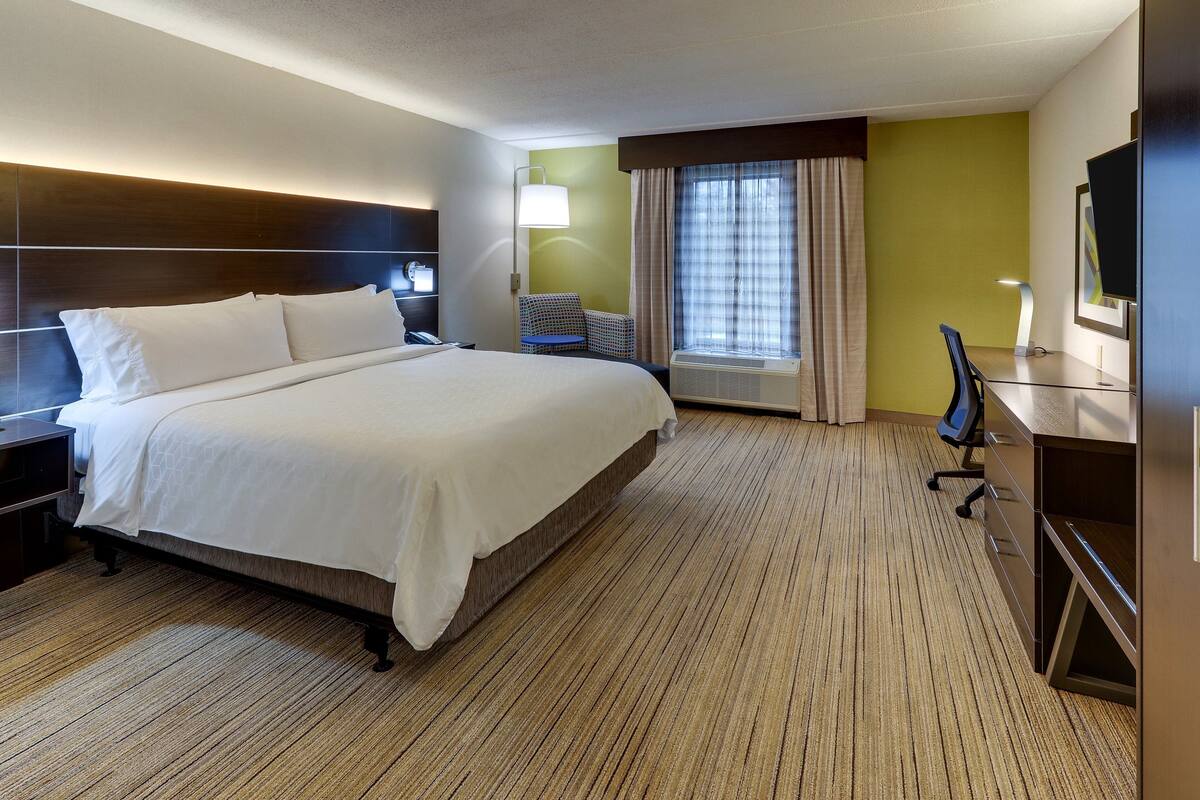 A hotel room with a large bed, a desk, a chair, a TV, and an air conditioning unit.