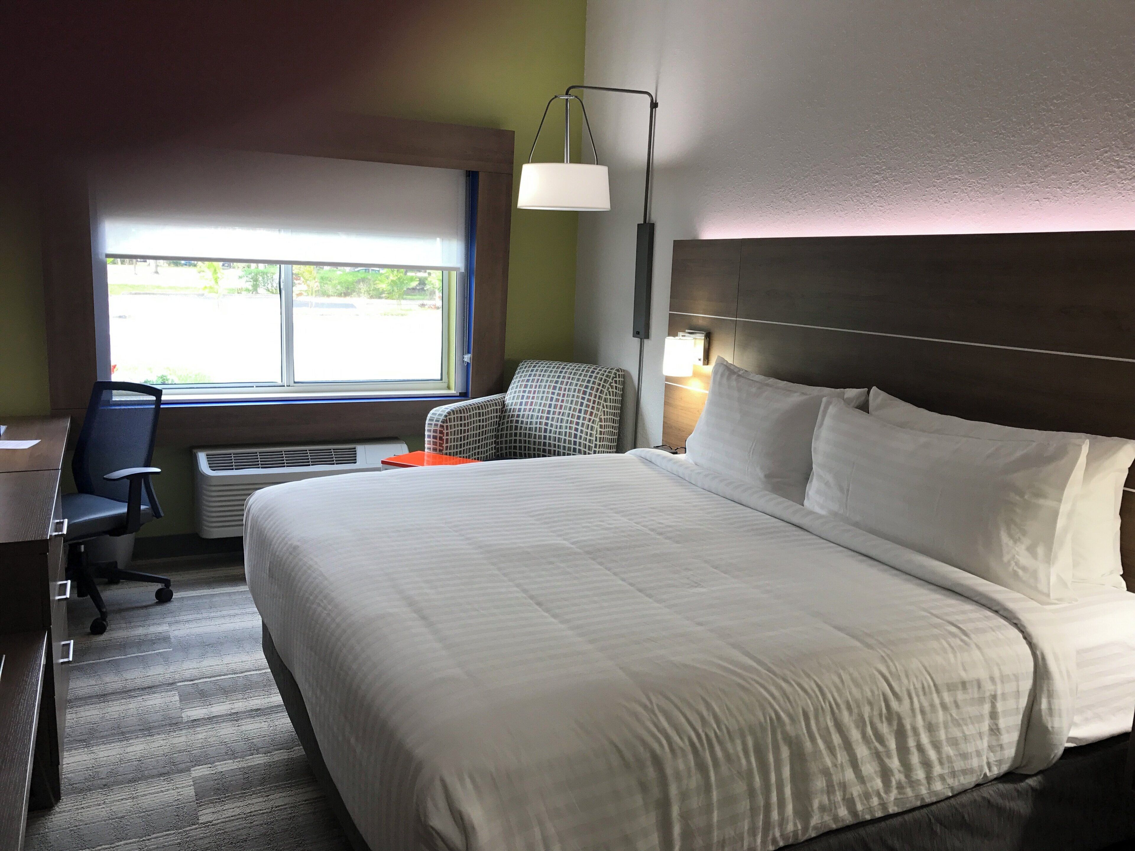 Une chambre d’hôtel avec un grand lit, un bureau, une chaise de bureau, une fenêtre avec des stores et une lampe fixée au mur.