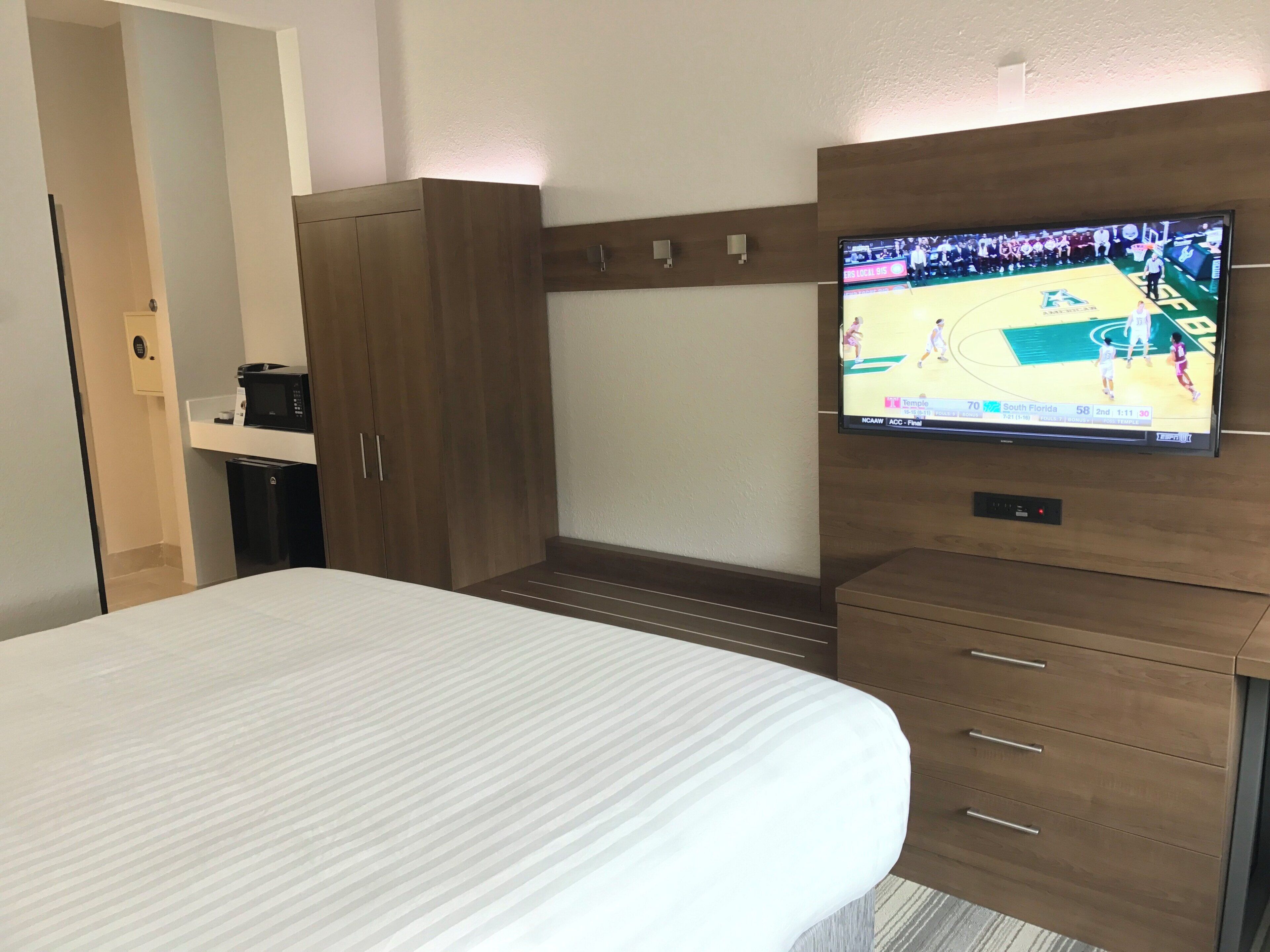 A hotel room with a bed, a television mounted on the wall, a wardrobe, and a small desk with a coffee machine.