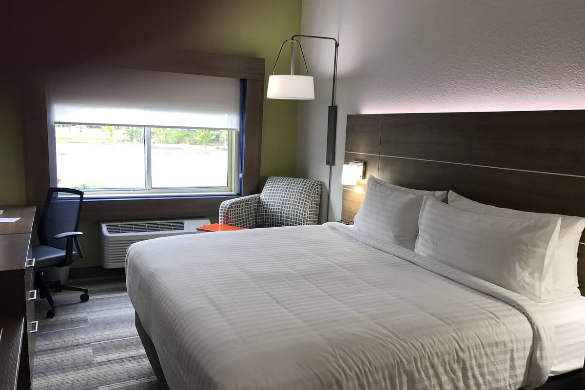 A hotel room with a large bed, a desk, a chair, a window with blinds, and a wall-mounted lamp.
