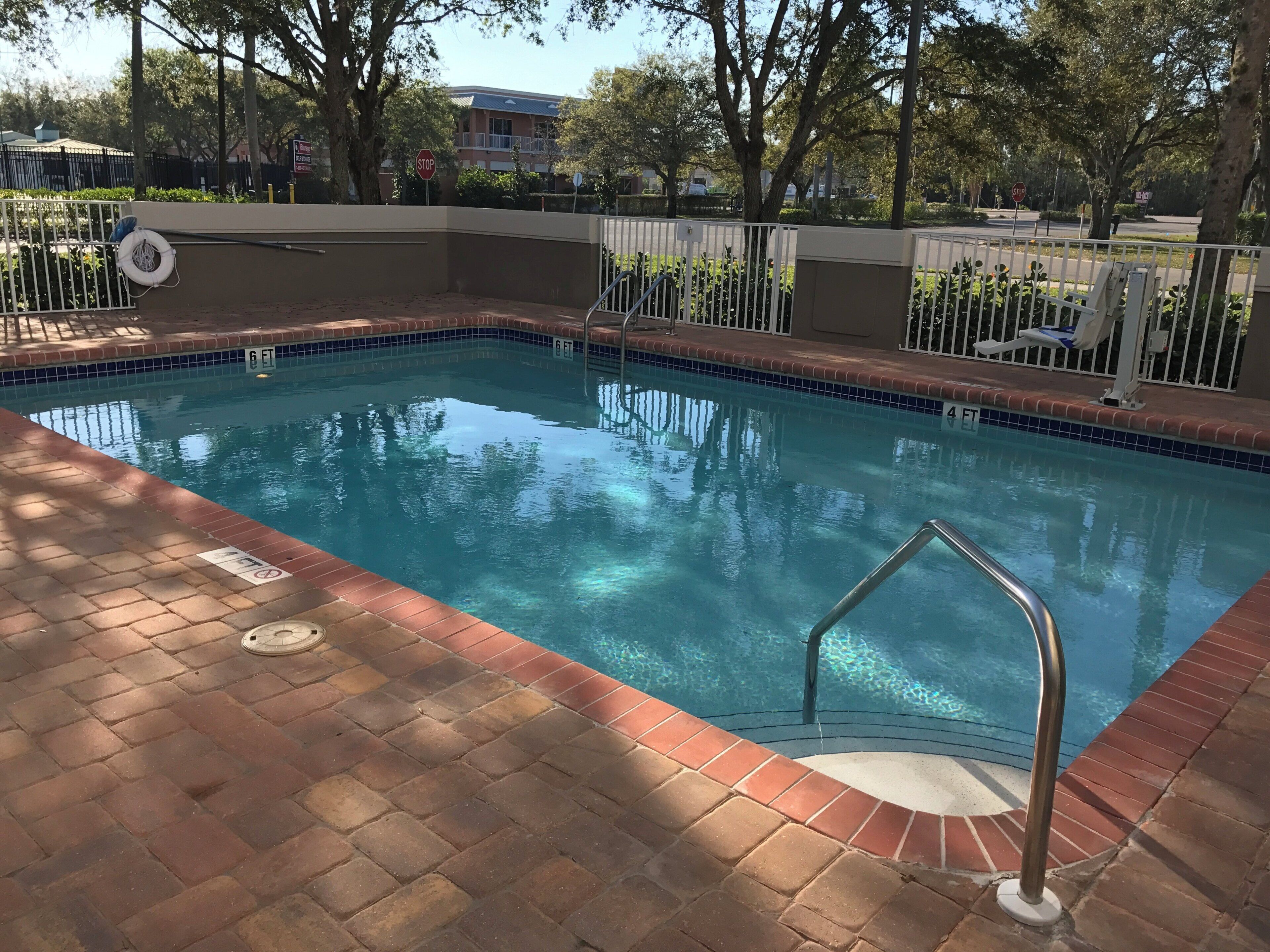 Una piscina con barandilla metálica de seguridad, rodeada por un patio de ladrillo y una valla metálica.