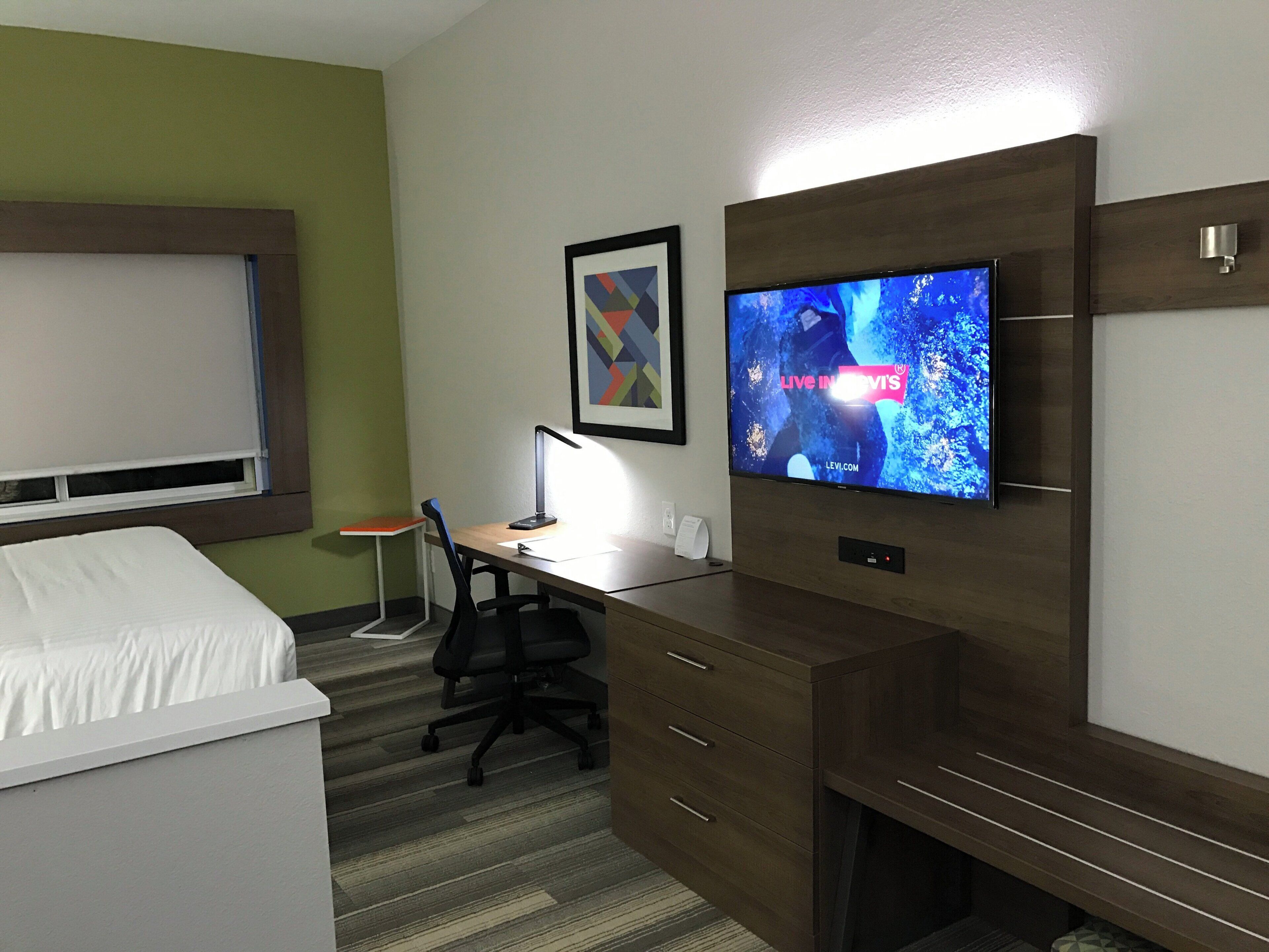 A hotel room with a bed, a desk with a computer, a chair, a television on the wall, and a framed picture.