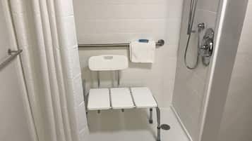 A small, white, tiled bathroom with a shower, a toilet, and a bench.