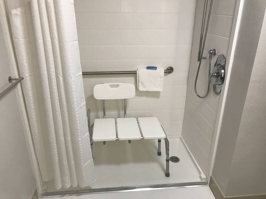 A small, white, tiled bathroom with a shower, a toilet, and a bench.
