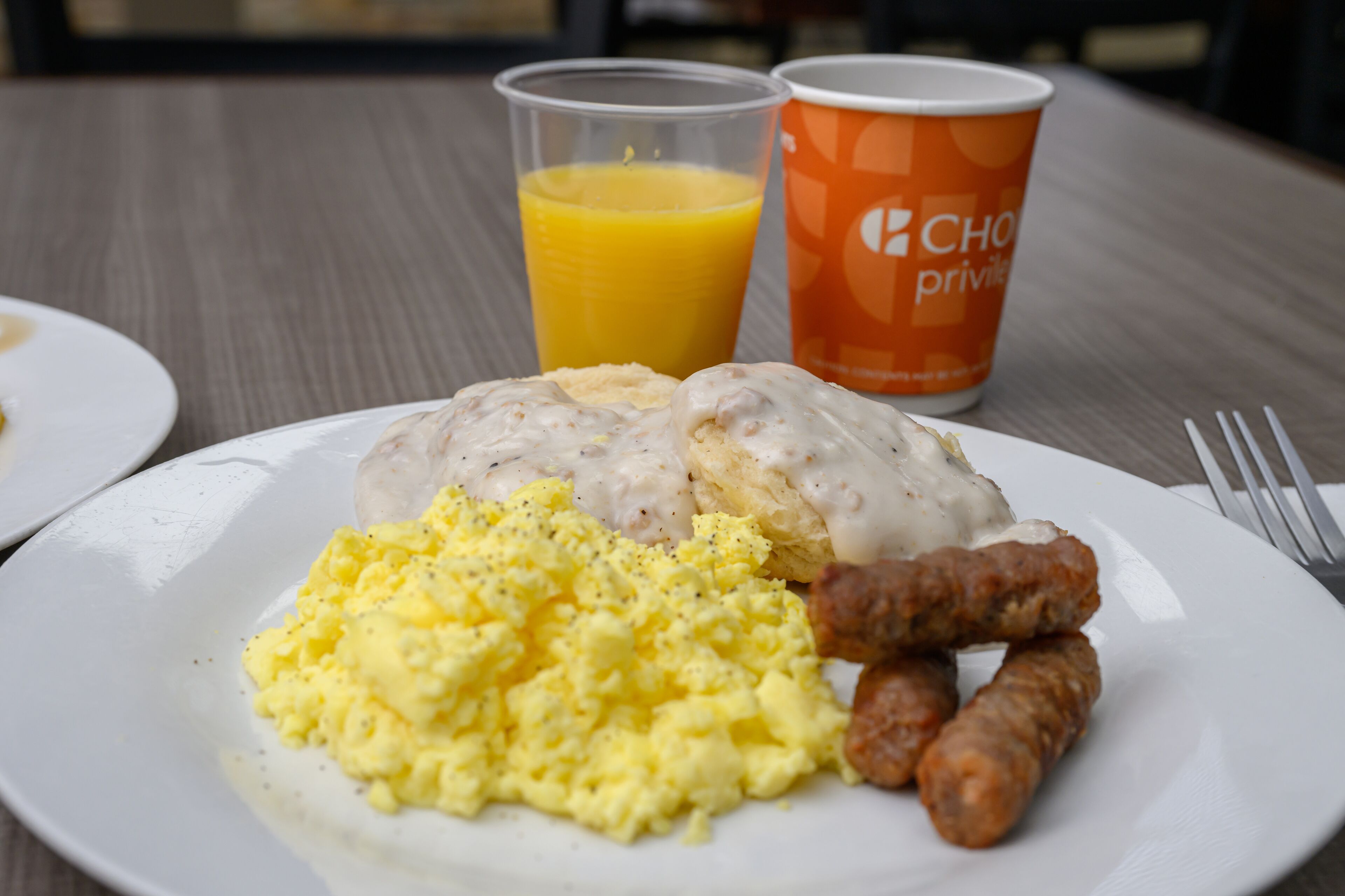 Un plato de desayuno con huevos revueltos, salchicha y salsa, acompañado de un vaso de jugo de naranja y una taza de café con el logo de la marca.
