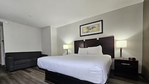 Una habitación de hotel con una cama grande, dos lámparas de noche, una mesita de noche, un sofá oscuro y un cuadro en la pared.
