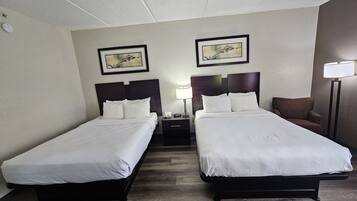 A hotel room with two single beds, a nightstand with a lamp, a chair, and two framed pictures on the wall.