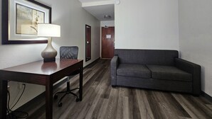 Une chambre d’hôtel avec un plancher en bois, un canapé gris, un bureau avec une lampe et un tableau encadré au mur.