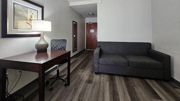 Quarto de hotel com piso de madeira, um sofá cinzento, uma escrivaninha com abajur e um quadro na parede.