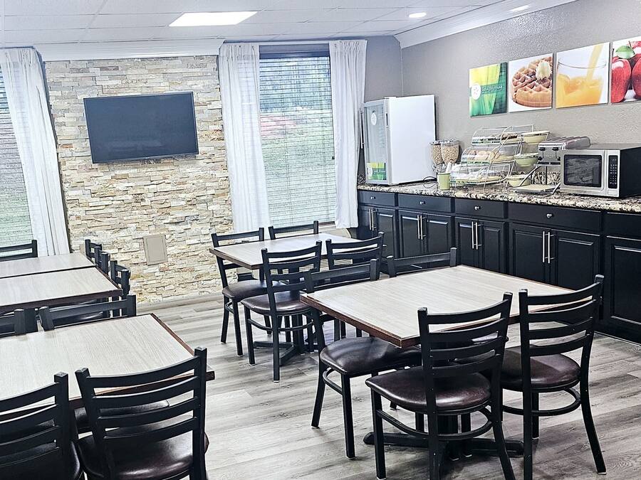 Un restaurante con pared de piedra, mesas de madera, sillas negras, una zona de cocina con electrodomésticos y un televisor montado en la pared.