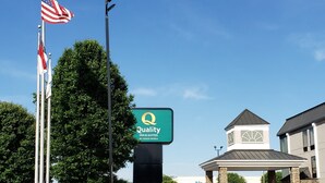 Entrada de um hotel com placa da Quality Inn & Suites by Choice Hotels, um mastro com a bandeira americana e um edifício com design moderno.