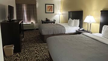 Habitación de hotel con dos camas, un escritorio, una silla, un televisor, una lámpara y una alfombra estampada.