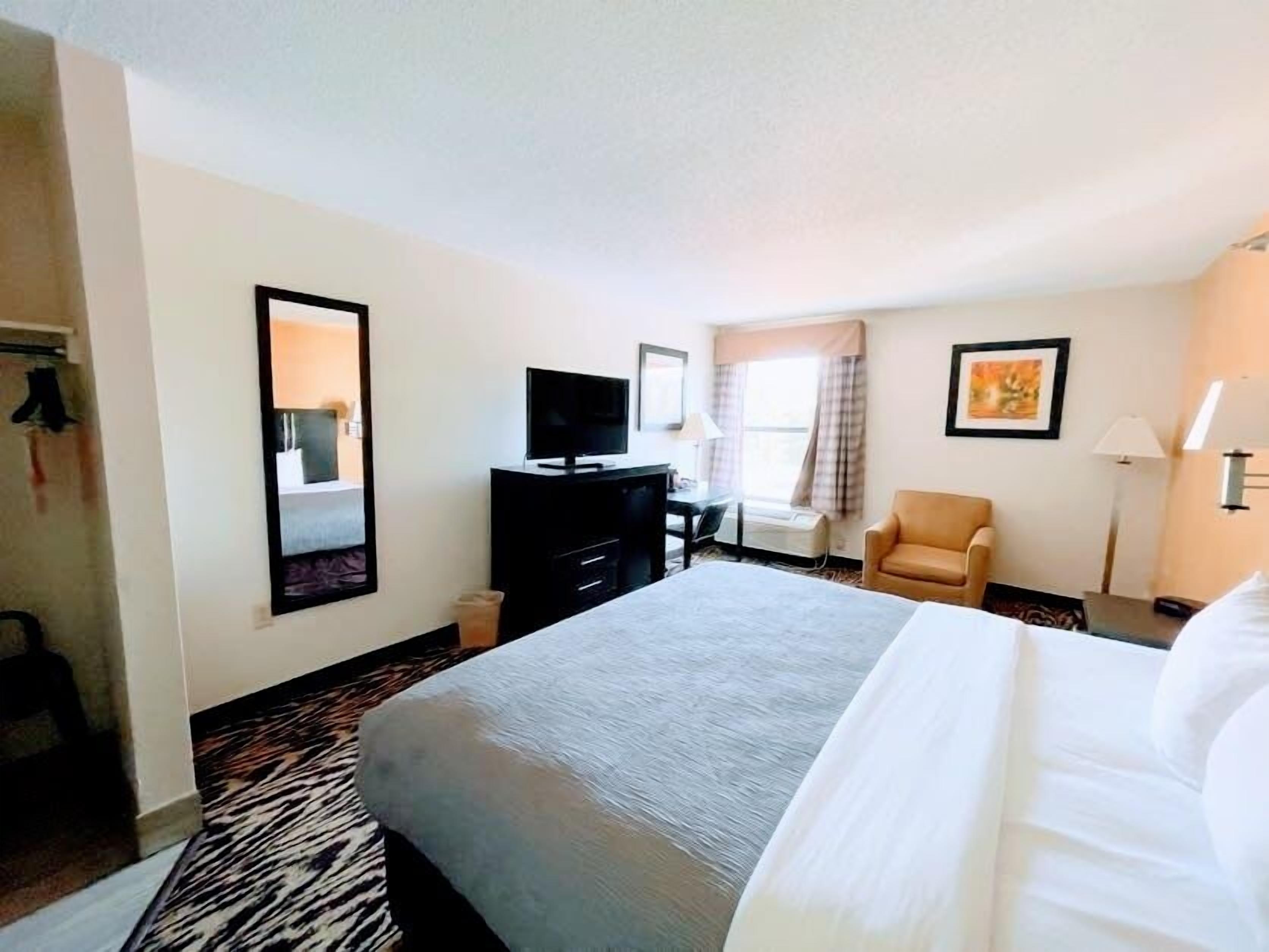 Quarto de hotel com uma cama grande, uma escrivaninha com computador, uma cadeira, televisão e janela com cortinas.